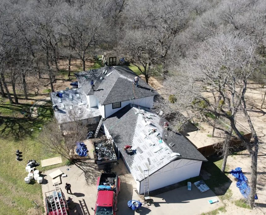 Aerial view of a house undergoing residential roofing with workers on the roof, surrounded by trees, a truck, and construction materials scattered around.