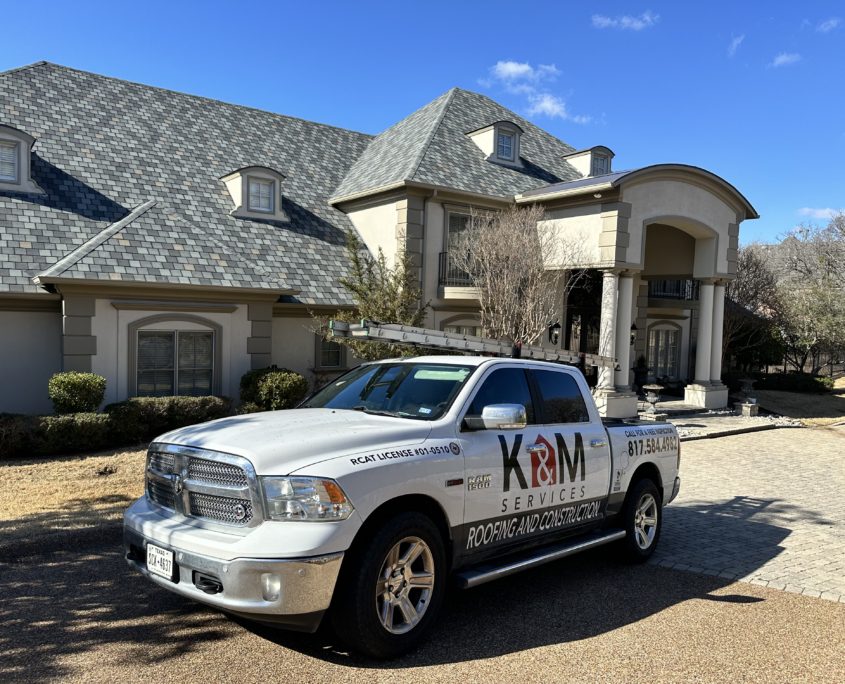 A white dodge ram truck branded with "K&M Services Residential Roofing Contractors" parked in front of a large, multi-story house on a sunny day.