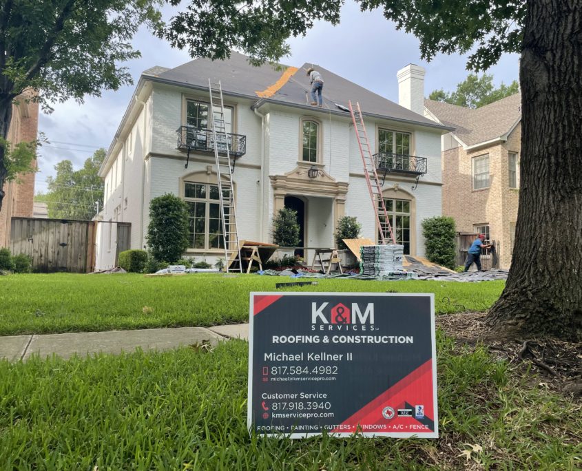 Workers repair the roof of a two-story residential house, with a lawn sign displaying roofing service contact details in the foreground.