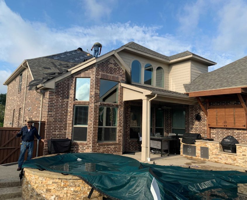 Two-story house with a man standing in the yard, a child near a fence, and residential roofing materials covering a section of the yard under a clear sky.