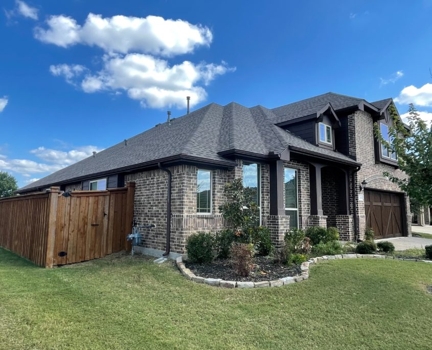 A modern two-story brick house with prominent residential roofing, manicured lawn, and an open wooden fence under a clear sky.