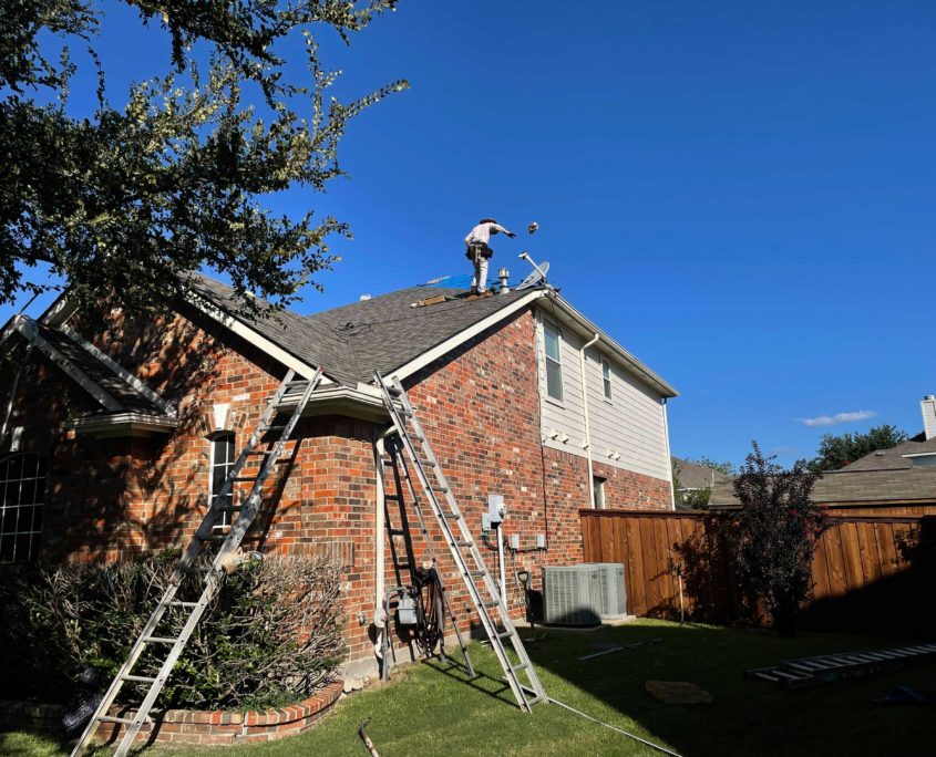 A person on a residential roofing inspecting or repairing shingles with a ladder propped against the building, under a clear blue sky.