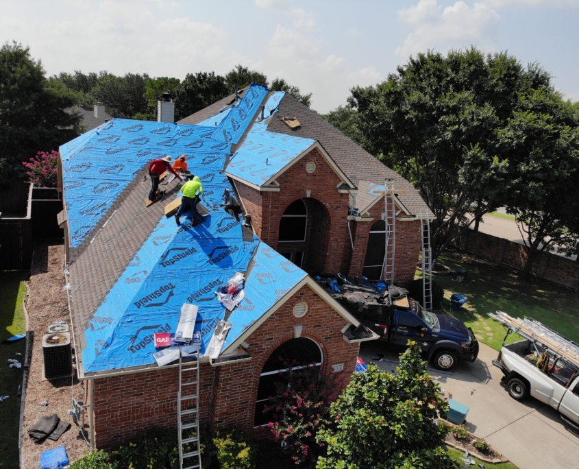 Aerial view of residential roofing workers replacing the roof of a two-story house, with blue protective sheeting covering the roof structure.