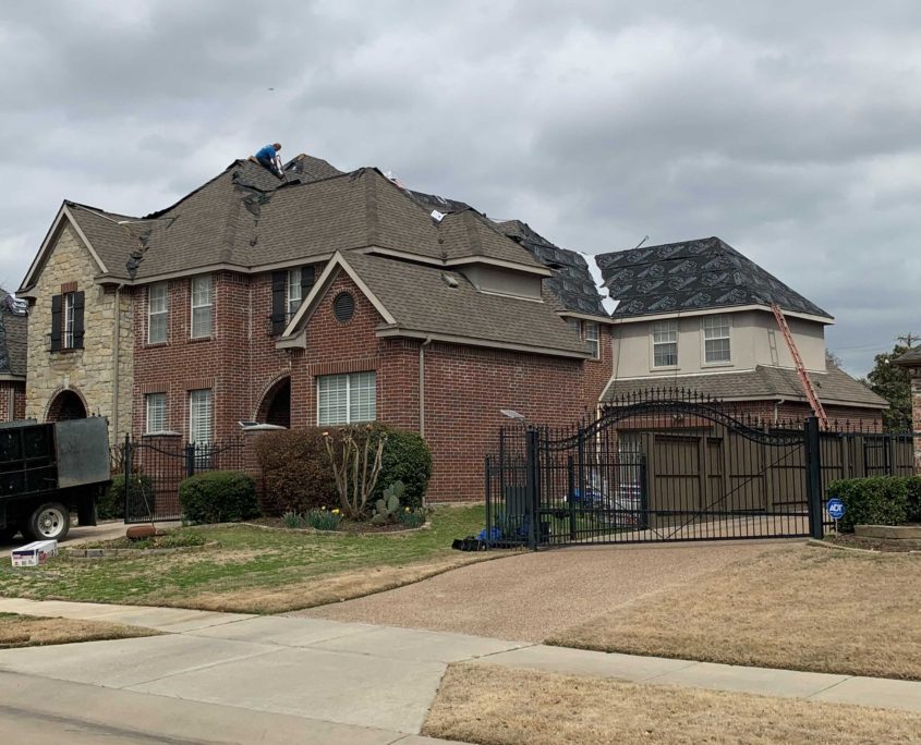 Workers on the roof of a large brick residential house, repairing damage under an overcast sky.