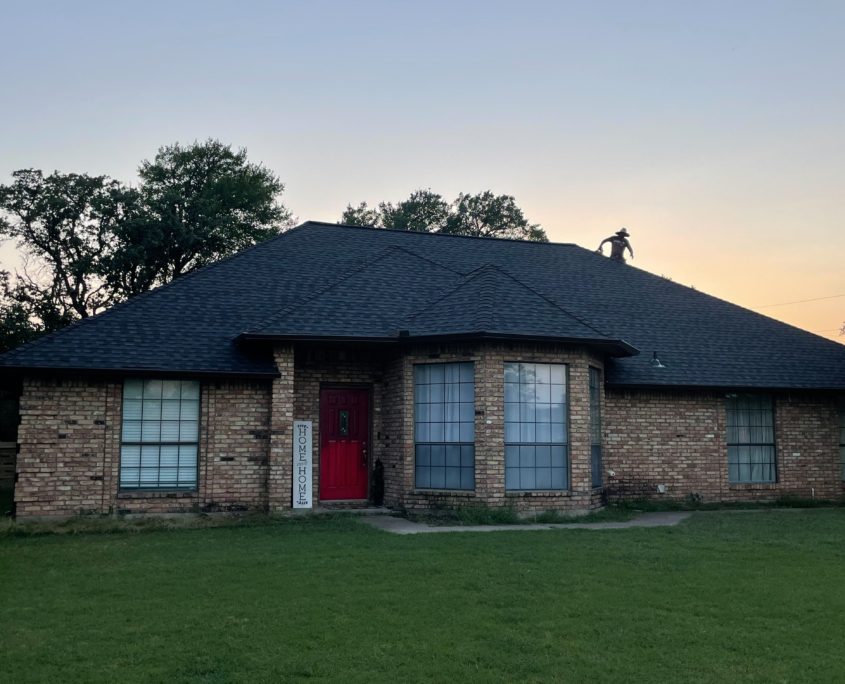 A single-story brick house with residential roofing and a red front door, surrounded by trees, at dusk. A person is visible on the roof.