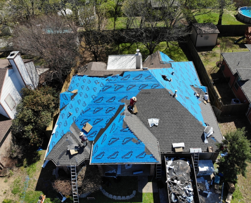 Aerial view of workers on a partially tarped residential roofing, with surrounding homes and greenery visible.